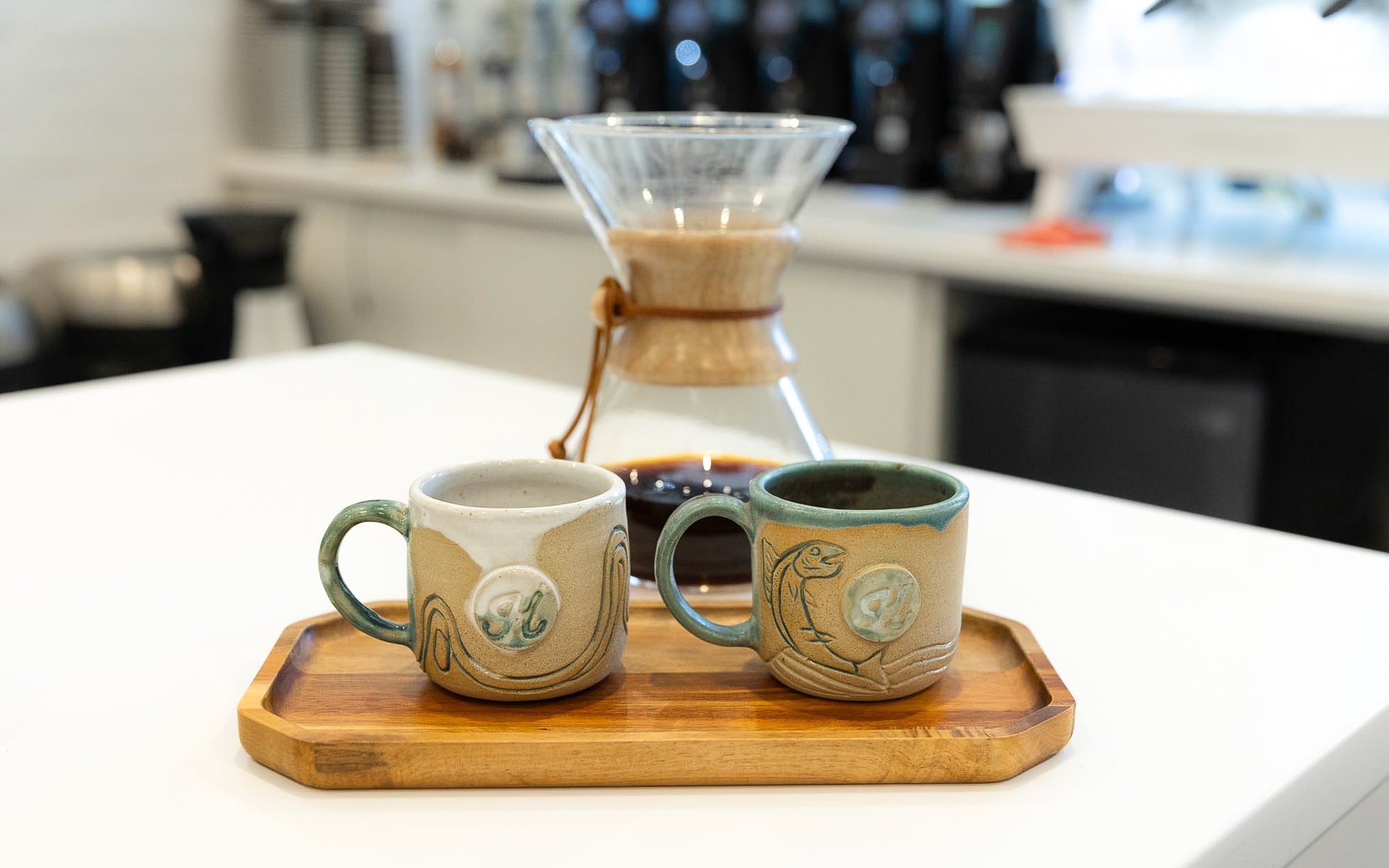 Two ceramic mugs on a wooden tray with a coffee brewing setup in a kitchen.