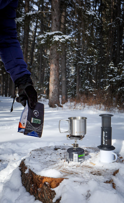 Coffee brewing equipment on a snow-covered log with a snowy forest background
