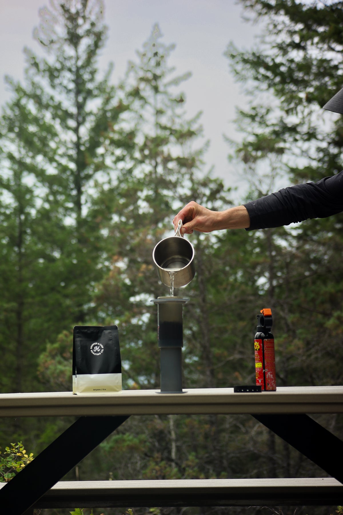 A person's arm is shown pouring hot water from a metal pitcher into a dark glass pour-over coffee maker set on an outdoor picnic table, with a bag of coffee and a bottle of syrup in the background against a forest backdrop.