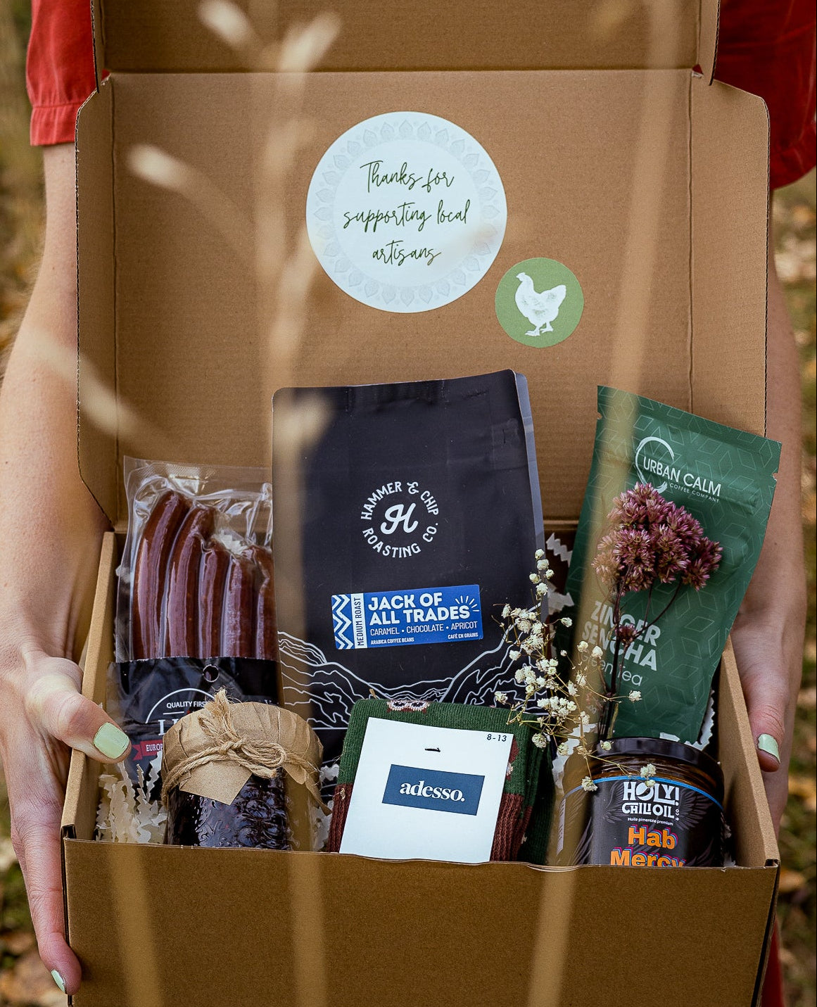 Person holding a box with various items including a blue bag and a green bottle, with a visible brand logo.