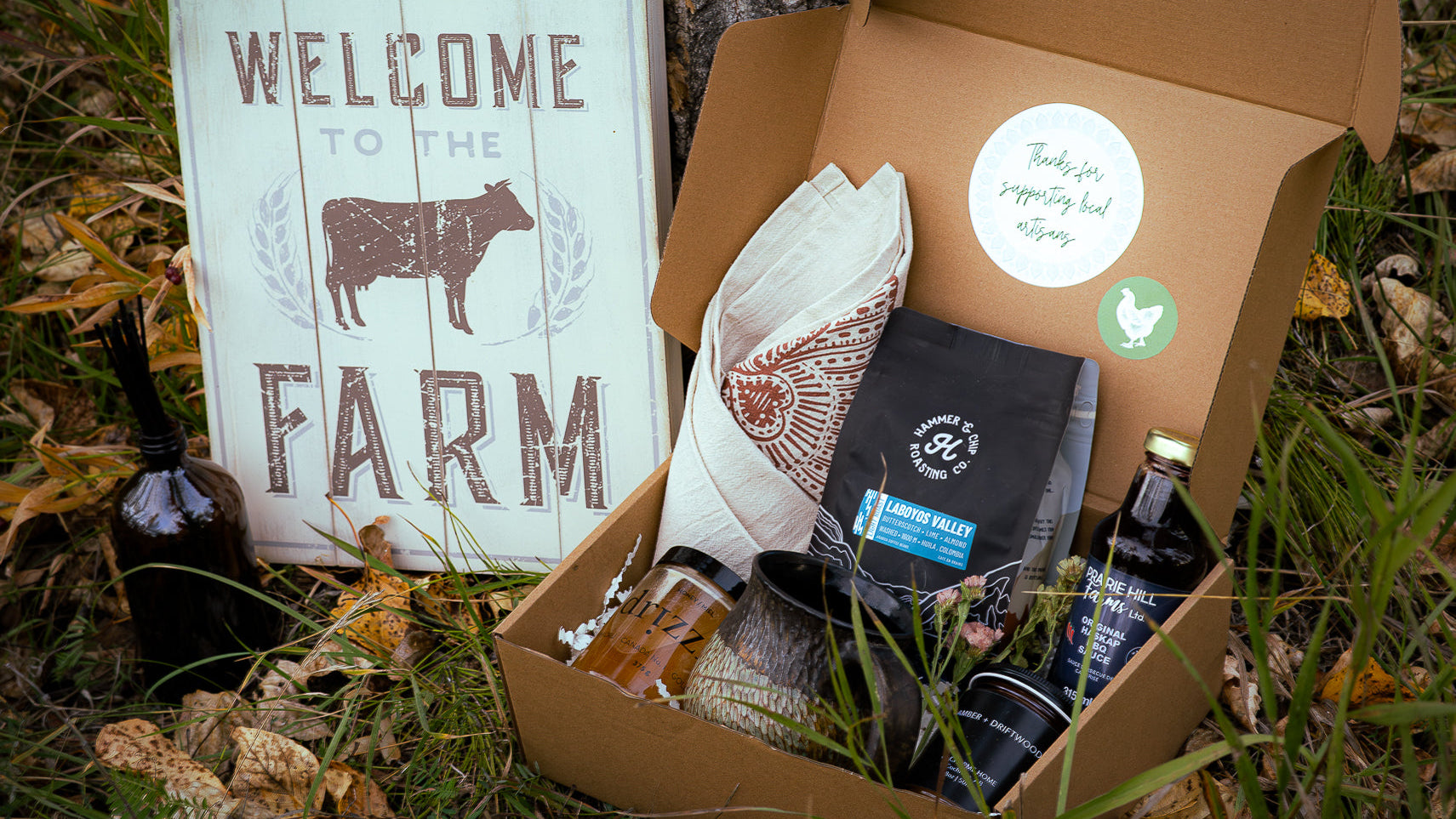 Box with farm-themed items including a bottle, a coffee bag, and a sign in a natural setting.